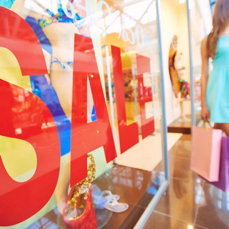 Vitrine de magasin avec le mot « soldes » et une jeune cliente portant des sacs en papier en arrière-plan.